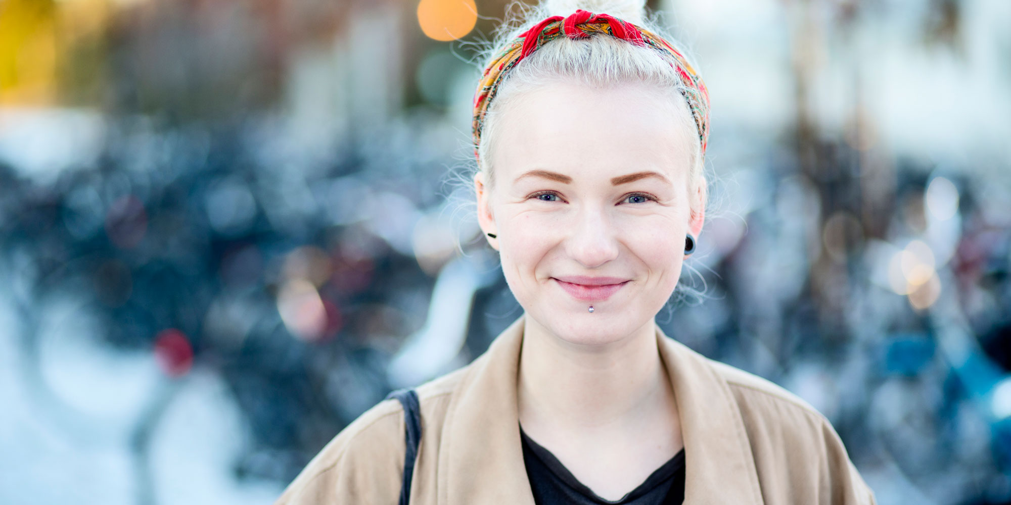 Ung blond leende kvinna fotograferad utomhus i uppsatt hår, med rött hårband och iklädd beige jacka.