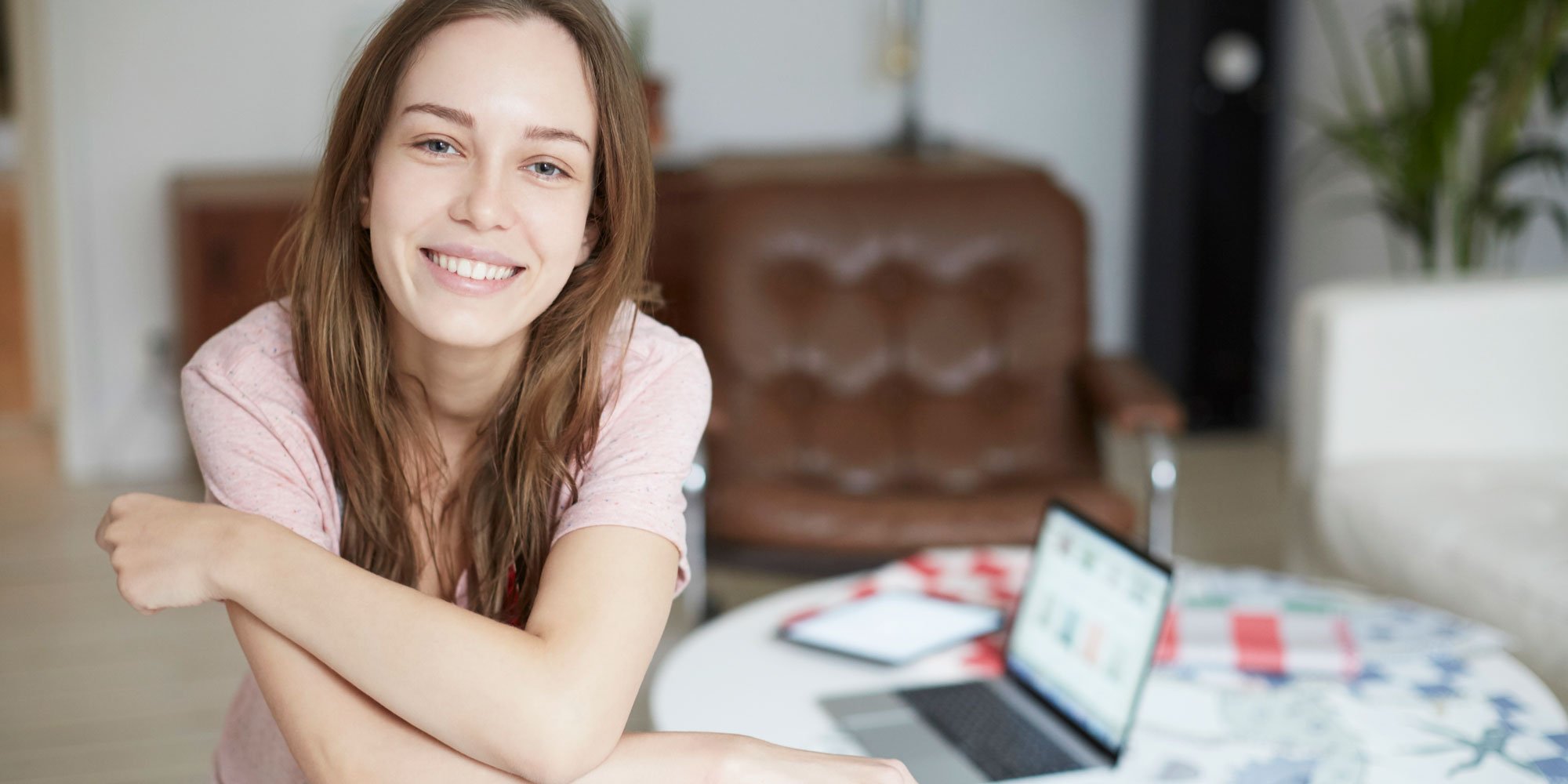 Leende ung kvinna i t-shirt sitter i hemmiljö vid ett lågt bord med en dator på bordet.