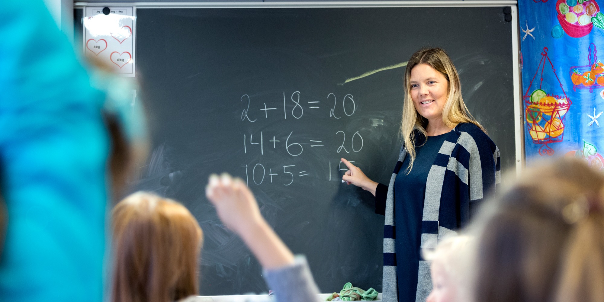 En kvinnlig lärare står framför en griffeltavla med mattetal på. Barn som håller upp händerna syns i förgrunden.