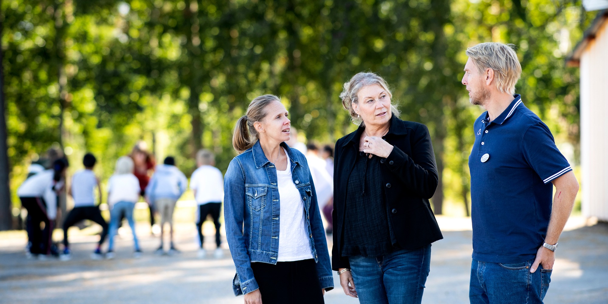 Tre personer står och pratar utomhus. I bakgrunden syns en skolgård med elever.