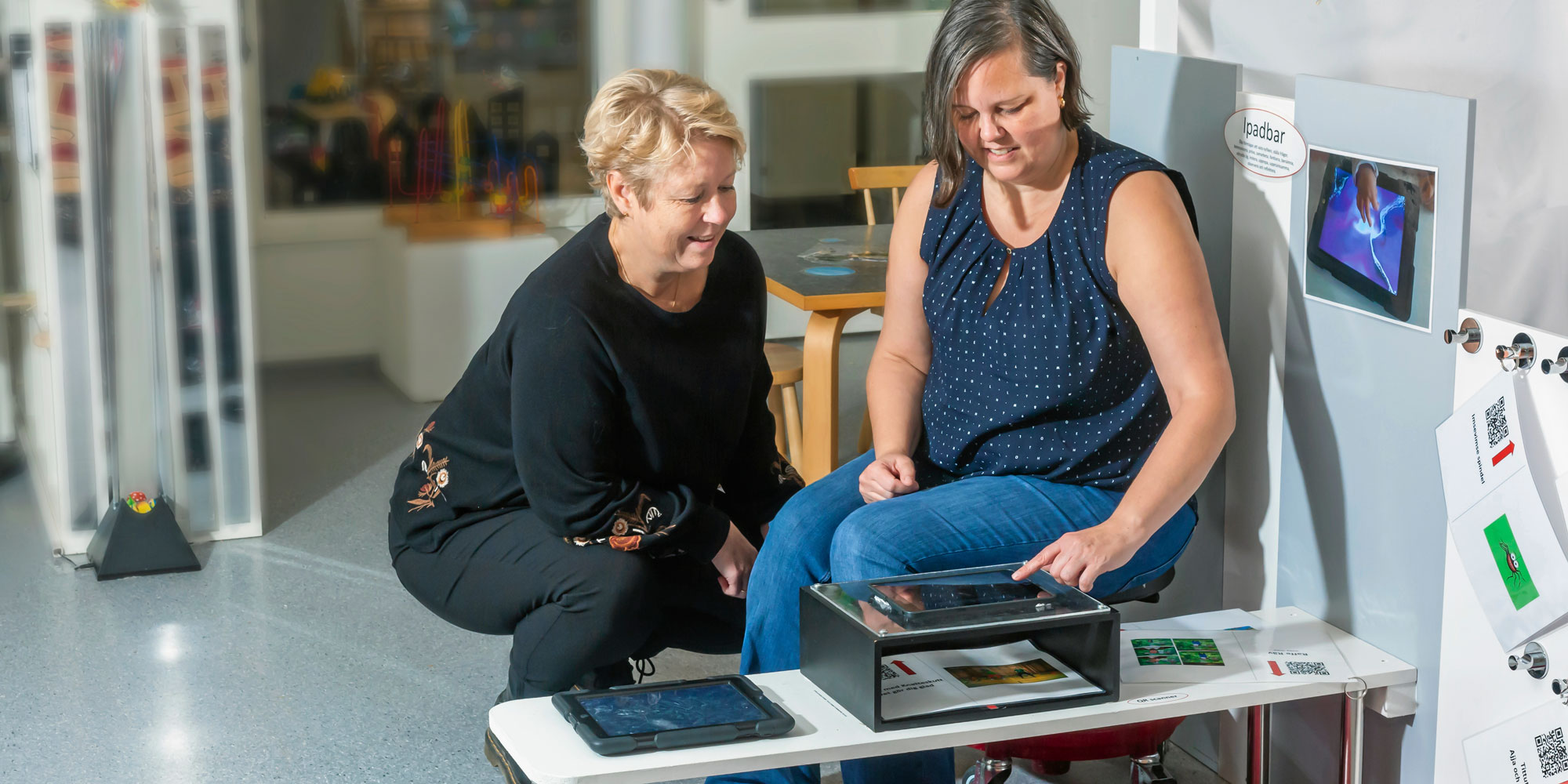 Förskolerektor Åsa Korén och barnskötare Ann-margreth Brandell sitter vid en litet bod med en Ipad framför sig.