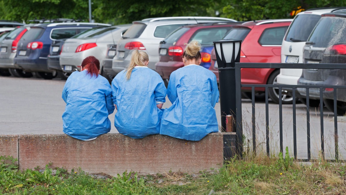 Tre personer i blåa skyddsrockar sitter med ryggen mot kameran på en mur vid en bilparkering.