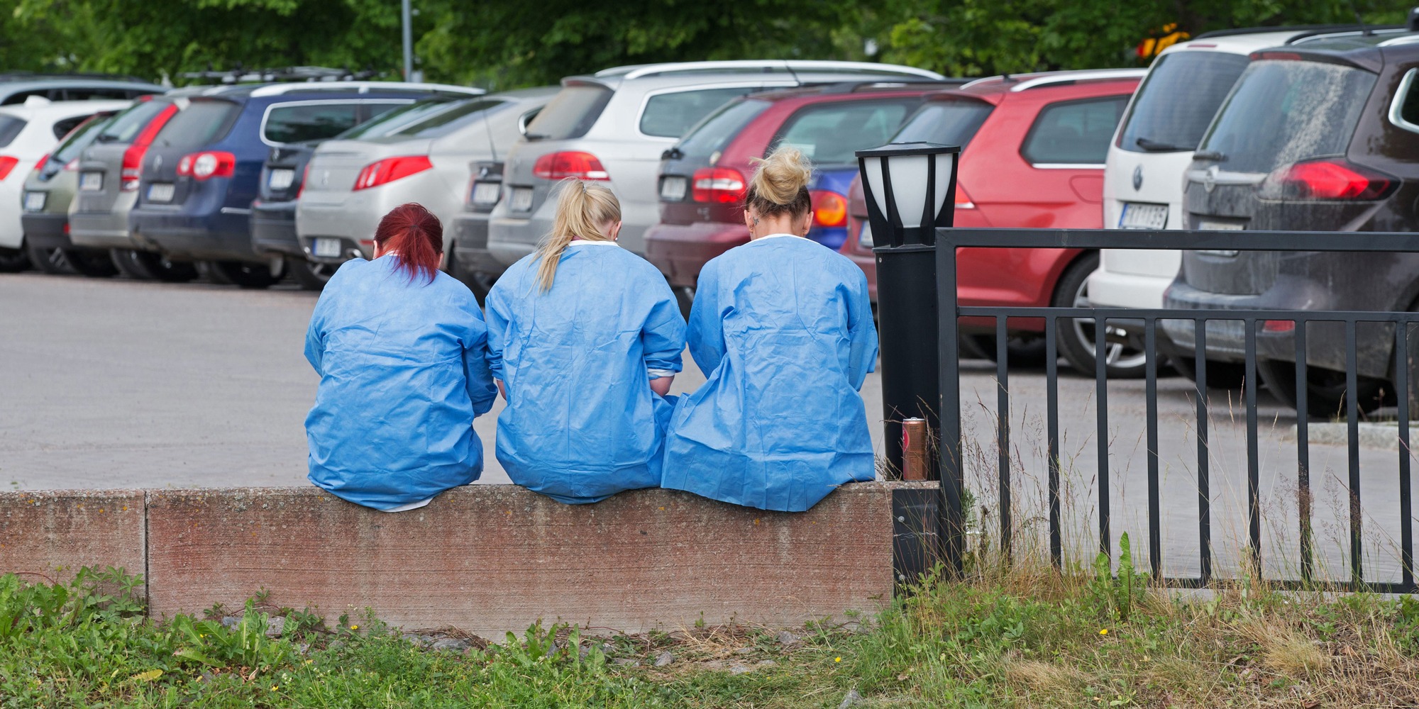 Tre personer i blåa skyddsrockar sitter med ryggen mot kameran på en mur vid en bilparkering.