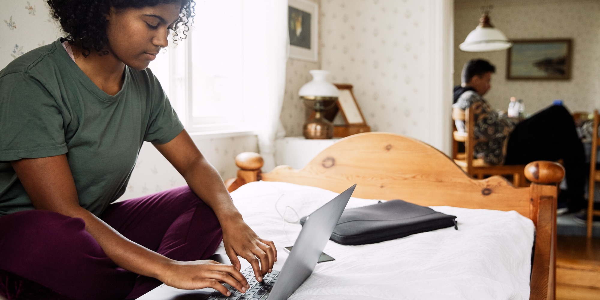 Ung kvinna sitter på säng med laptop framför sig.