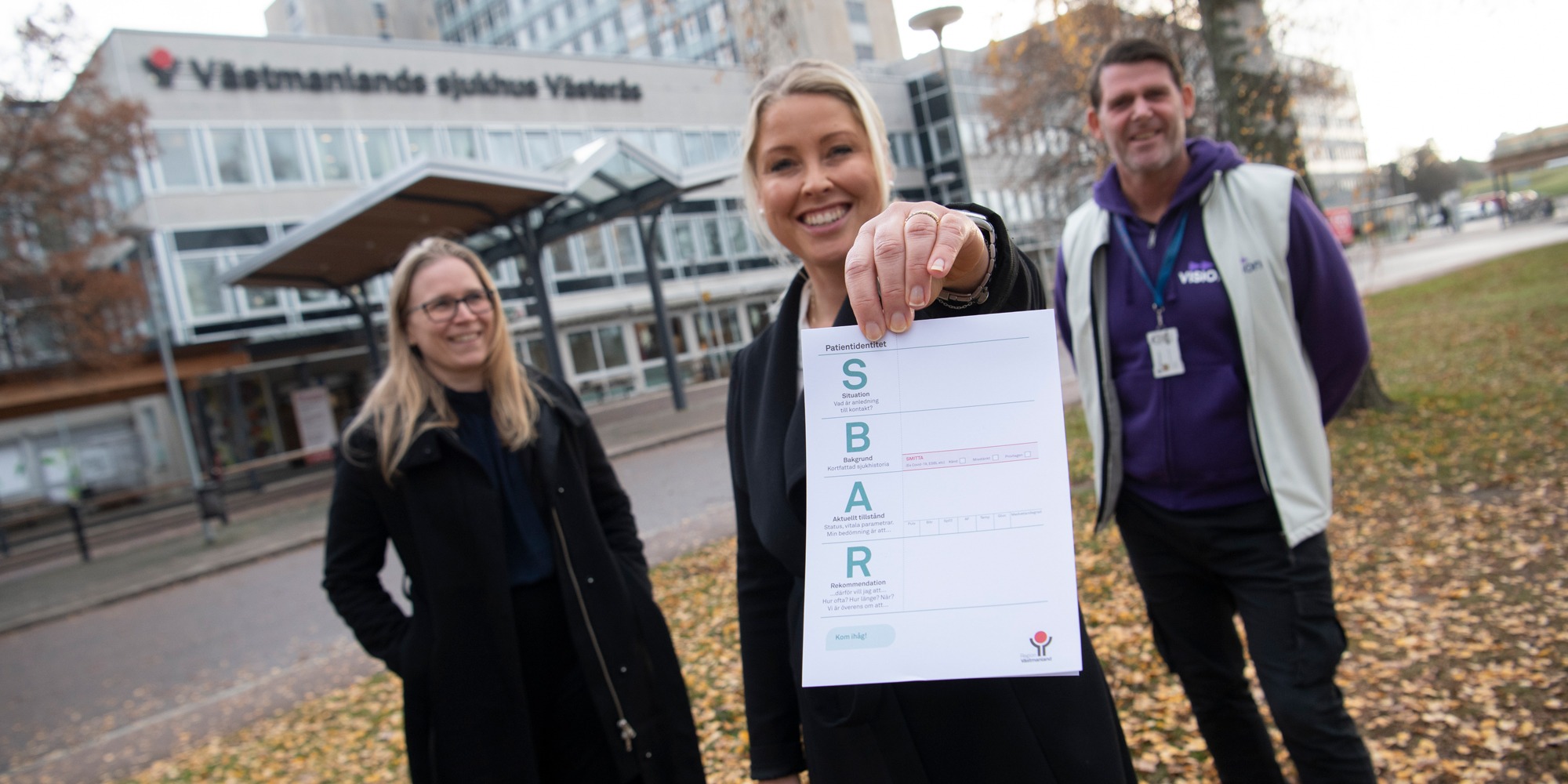 Tre personer står framför en sjukhusbyggnad. Person i mitten håller upp ett papper.