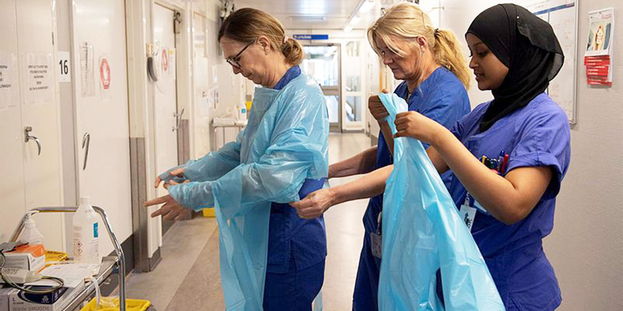 Tre kvinnor i en sjukhuskorridor som tar på sig skyddsrutrustning.