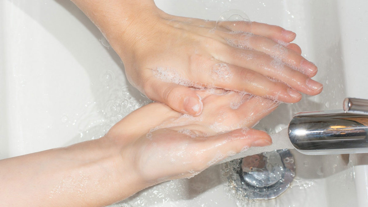 Två ljusa händer med tvål under rinnande vatten i ett handfat.
