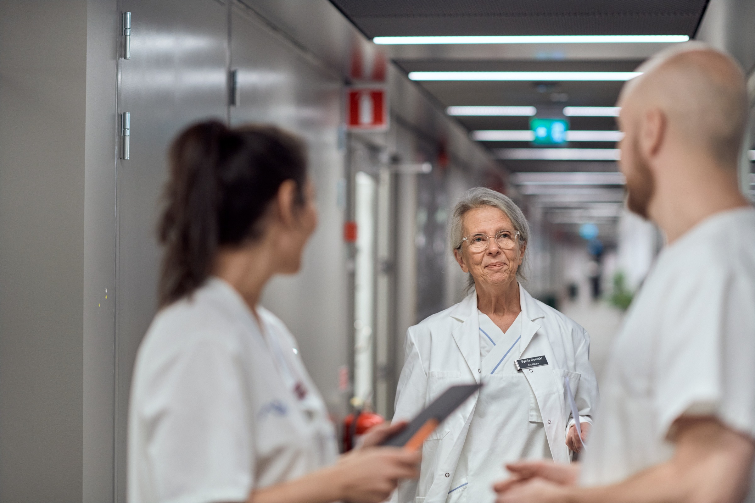 Kvinna i vita sjukhuskläder står i en korridor och samtalar med två kollegor.