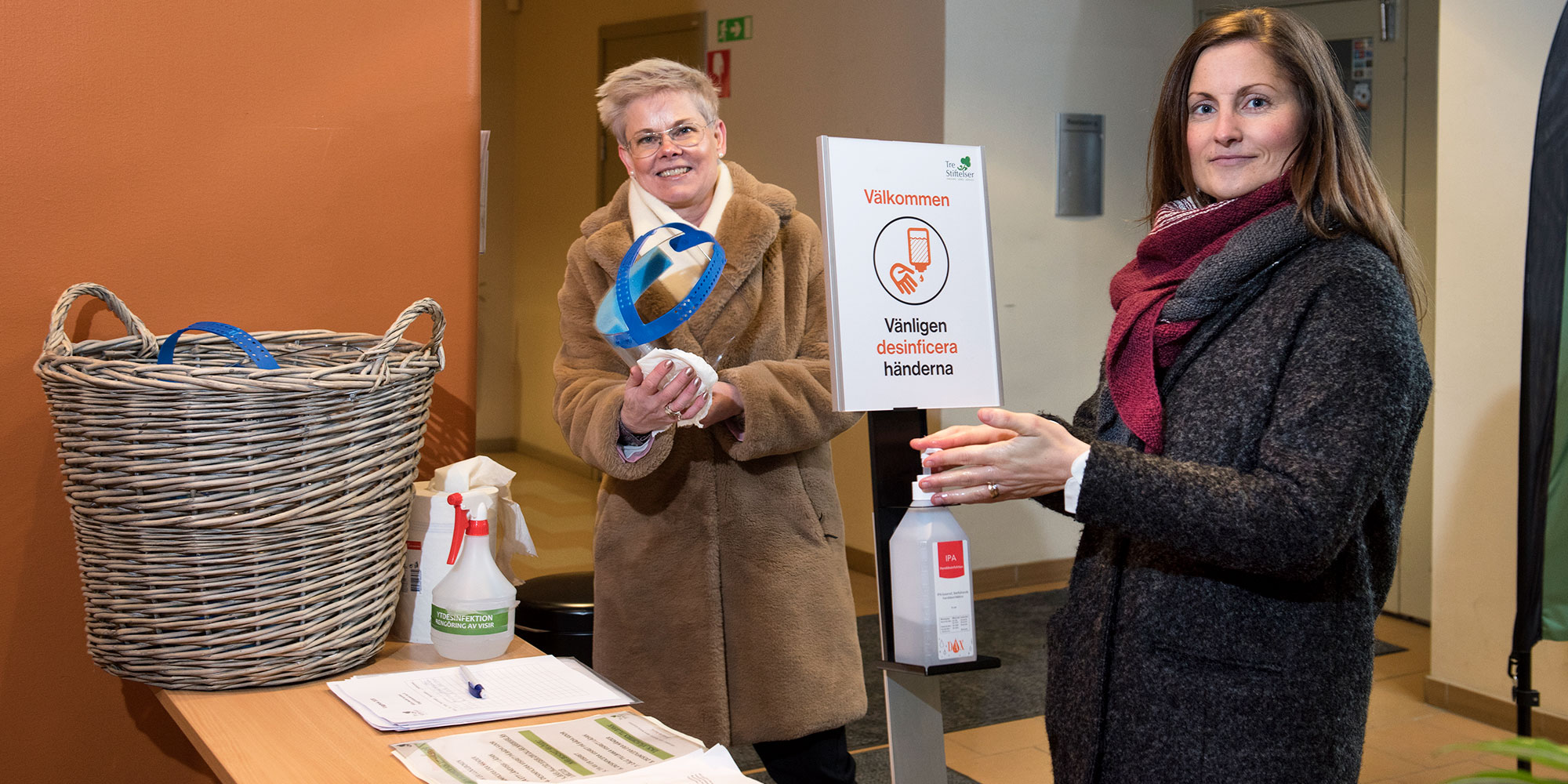 Två personer i ytterkläder står i entrén till ett äldreboende, med en pump för handsprit och en lista på ett bord där besökare skriver upp sig.