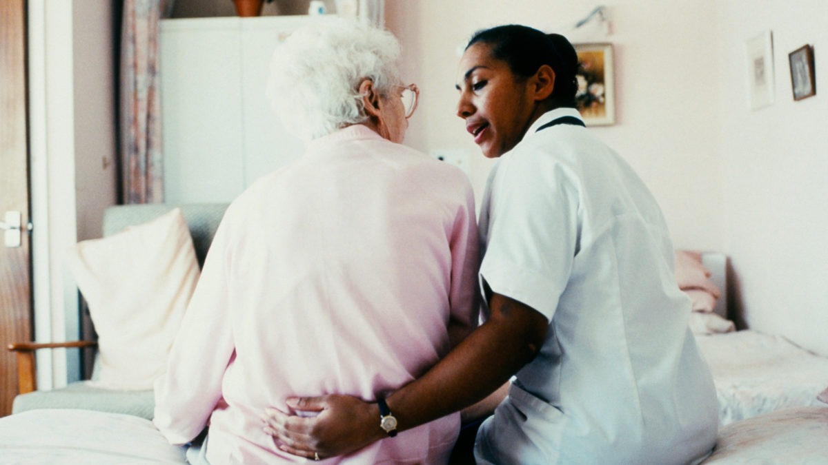 En äldre dam och en sköterska sitter på en säng