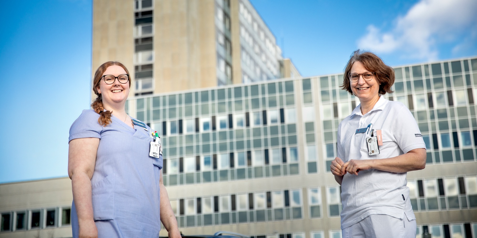 Två personer i sjukhuskläder står utomhus framför en sjukhusbyggnad. Blå himmel.