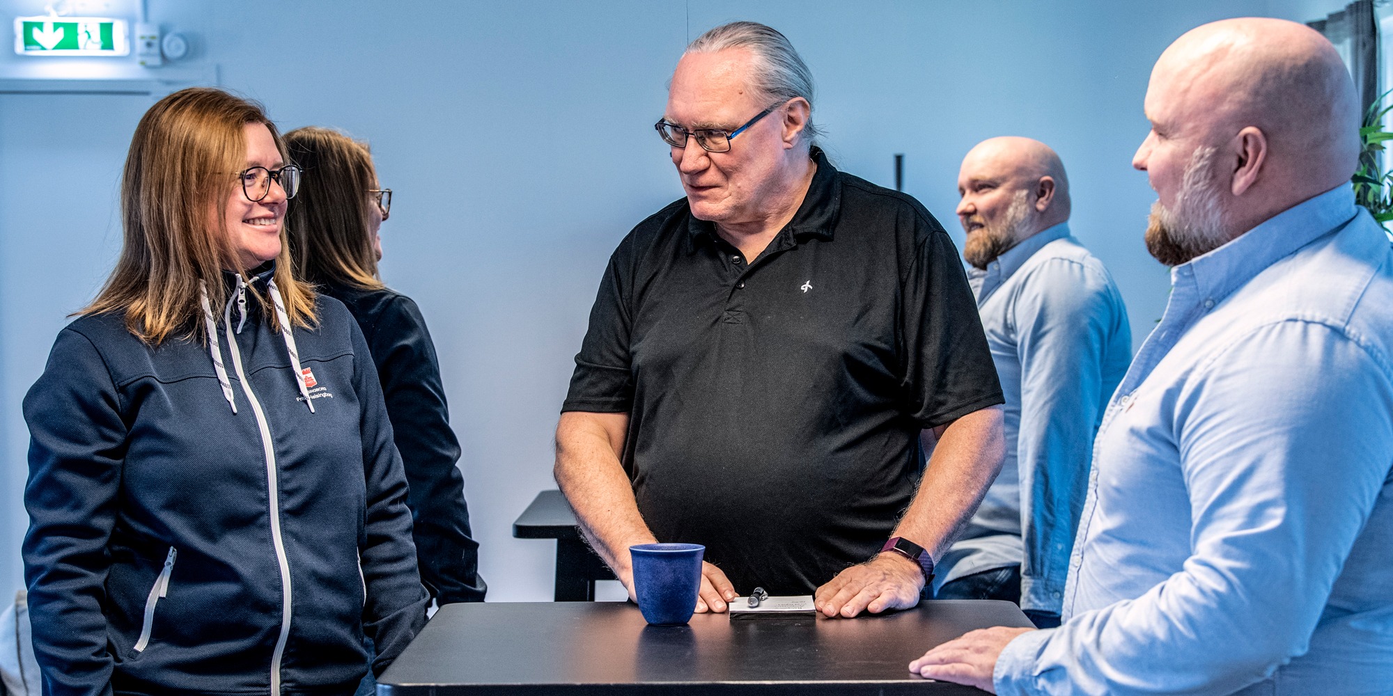 En grupp människor står och pratar vid ett bord. I bakgrunden en gigantisk spegel.