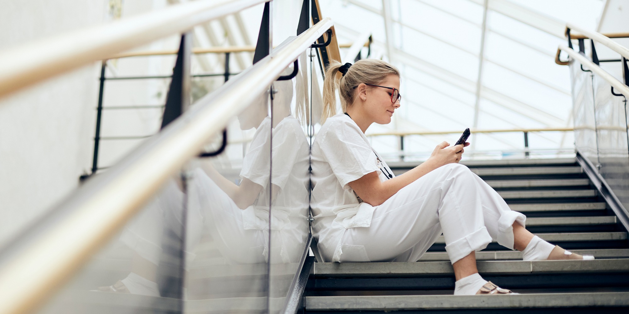 Kvinna i sjukhuskläder sitter i en trappa med sin mobiltelefon.