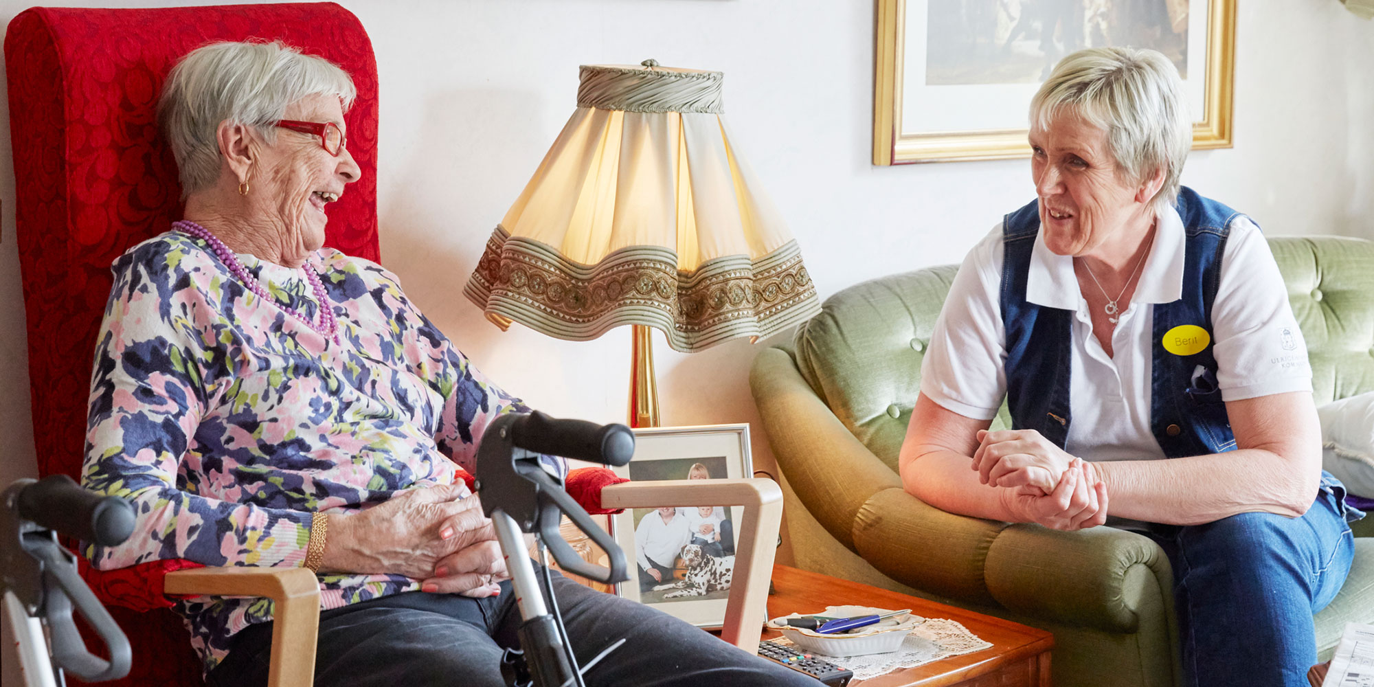 Äldre kvinna i fåtölj samspråkar med kvinna bredvid i soffa.