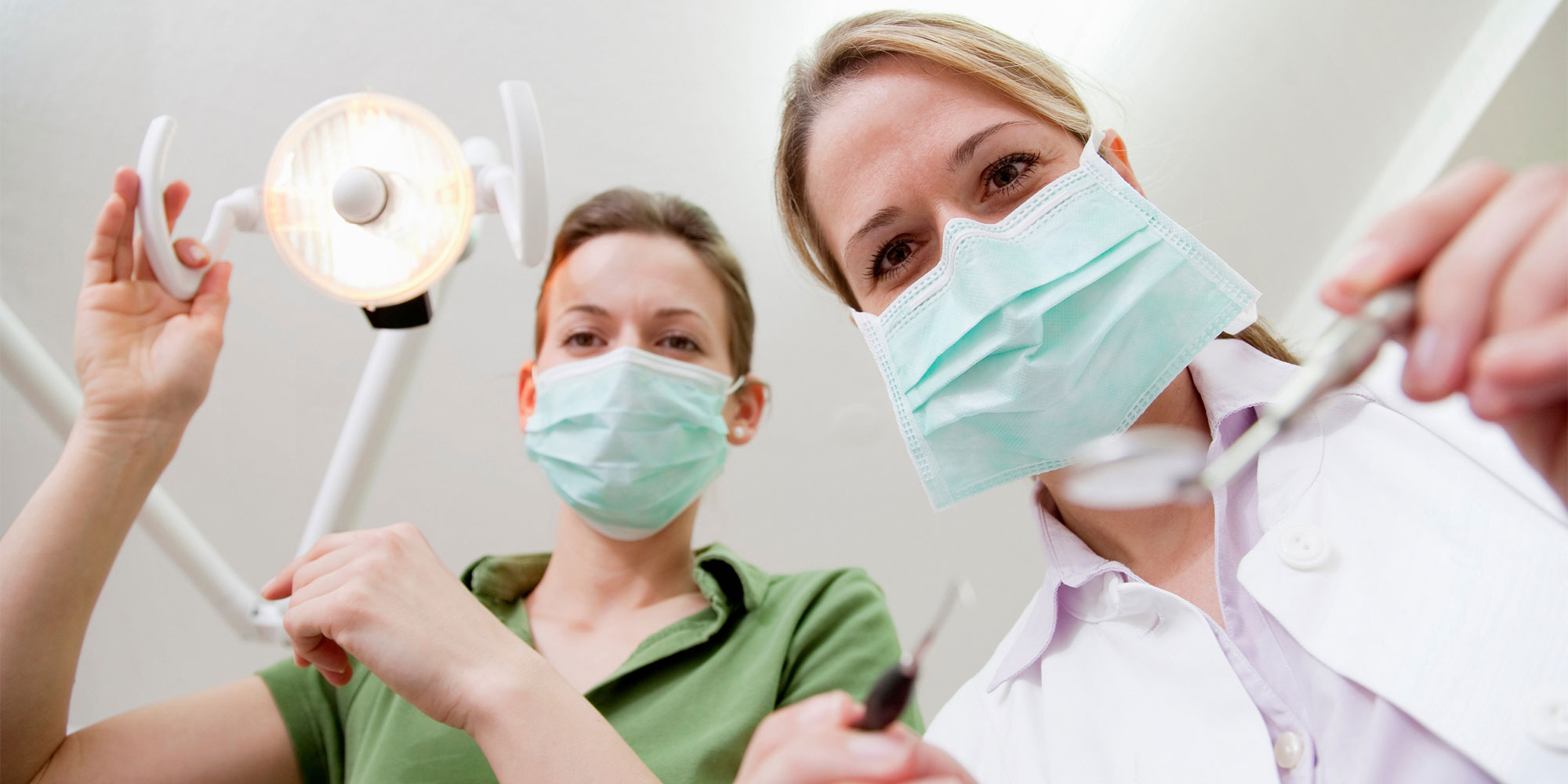 Två kvinnor i munskydd och vårdkläder lutar sig ner över betraktaren som verkar sitta i en tandläkarstol.