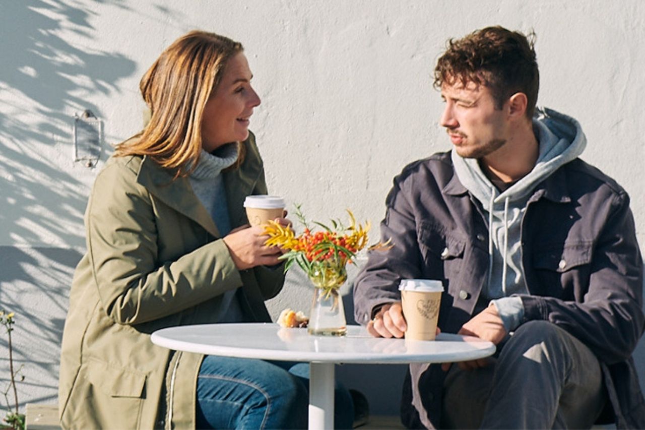 Två personer sitter vid ett cafébord utomhus och dricker kaffe och samtalar med varandra.