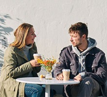 två personer sitter och fikar utomhus i solen, med jackorna på. 