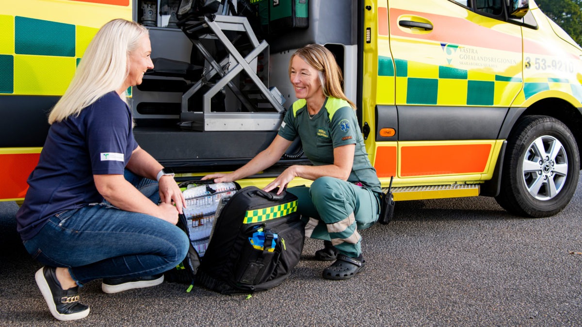 Två kvinnor sitter på huk och pratar framför en ambulans