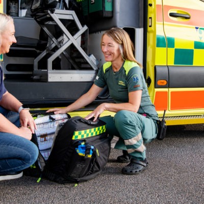 Två kvinnor sitter på huk och pratar framför en ambulans