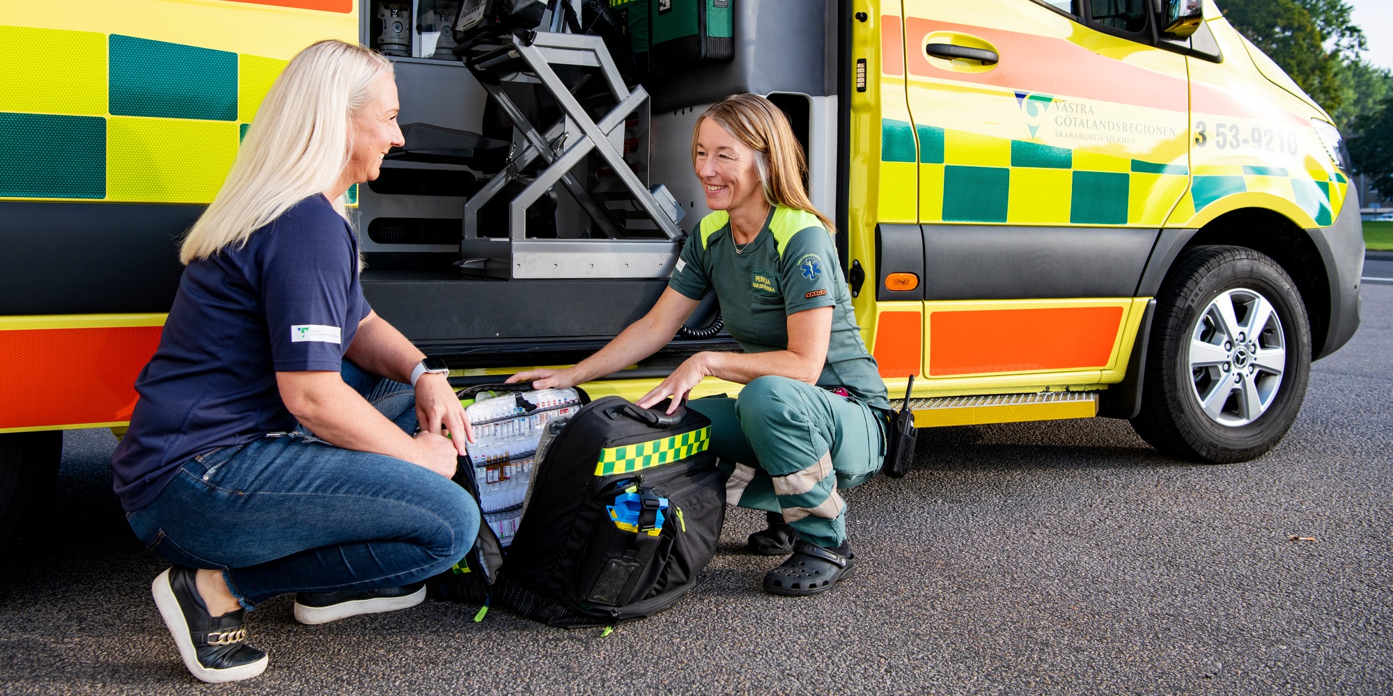 Två kvinnor sitter på huk och pratar framför en ambulans