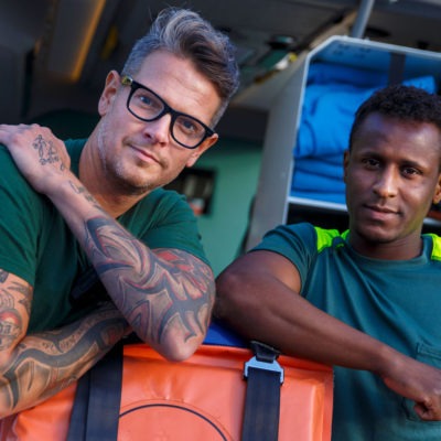 två män från räddningstjänsten utanför en ambulans. Tittar in i kameran.
