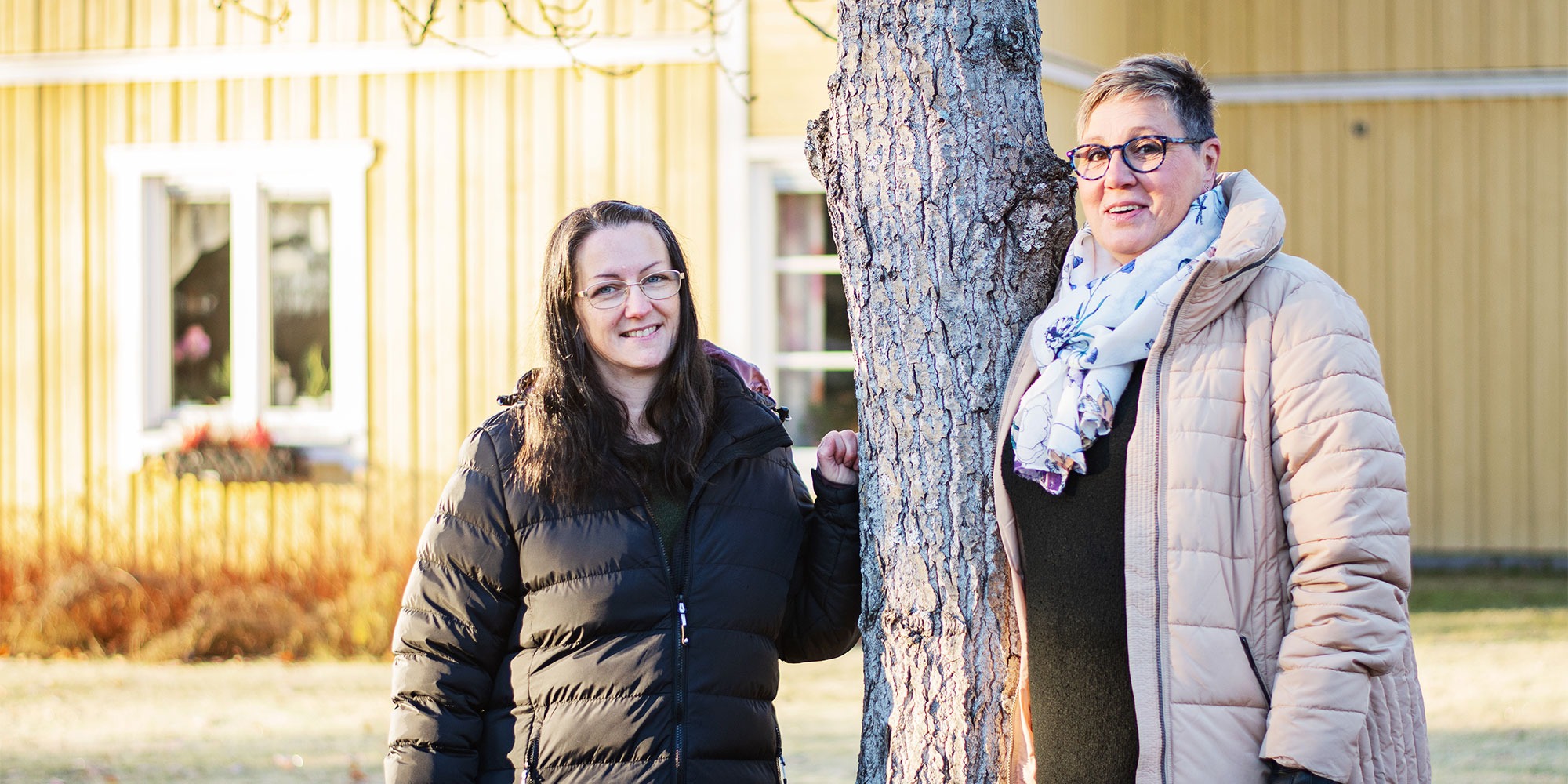 Skyddsombudet Petra Monwell och enhetschefen Madelen Karlsson vid et träd i vintersol utanför en gul vacker träbyggnad, Brismarksgården i Hoting, Strömsunds kommun.