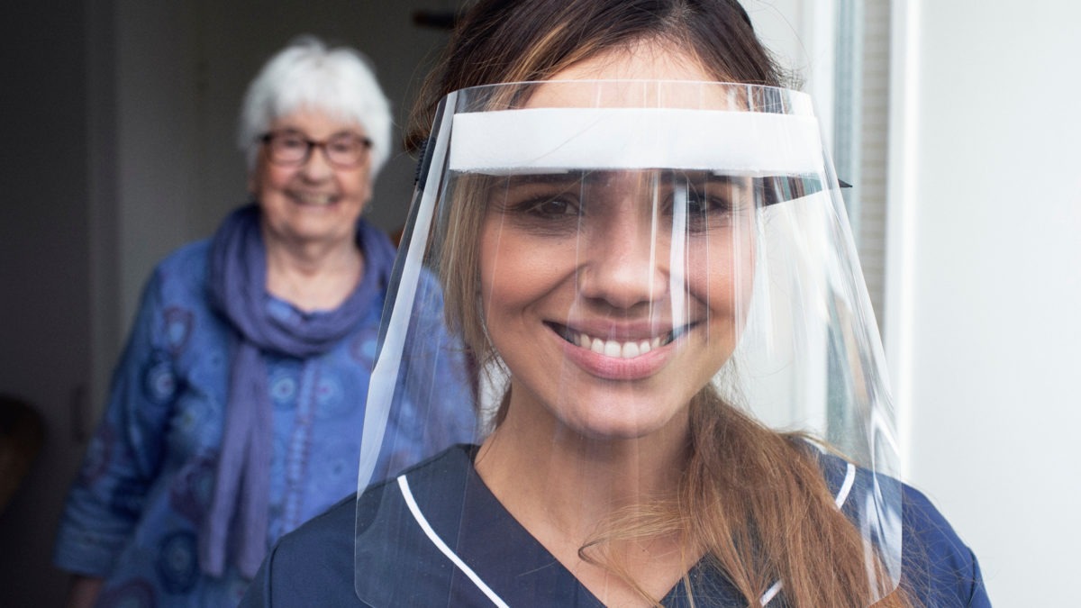 ung kvinna i vårdkläder och med visir ler mot kameran. I bakgrunden en äldre leende kvinna.