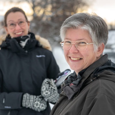 Två leende kvinnor står utomhus i snölandskap.