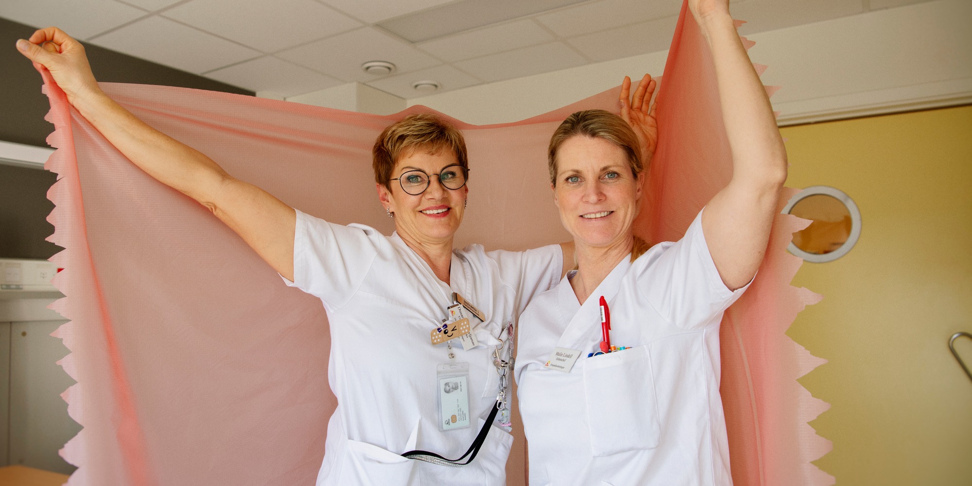 två kvinnor i vårdkläder håller upp ett så kallat glidlakan.