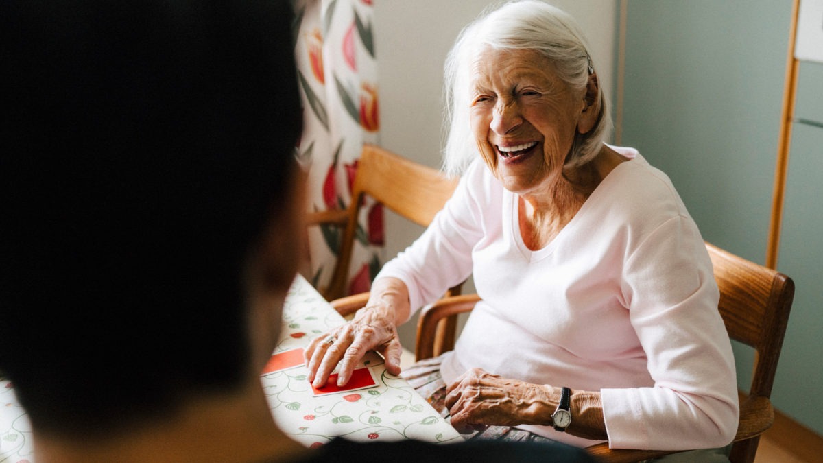 Äldre kvinna sitter ned vid bord och ler mot en ung hemtjänstanställd som står med ryggen mot kameran.