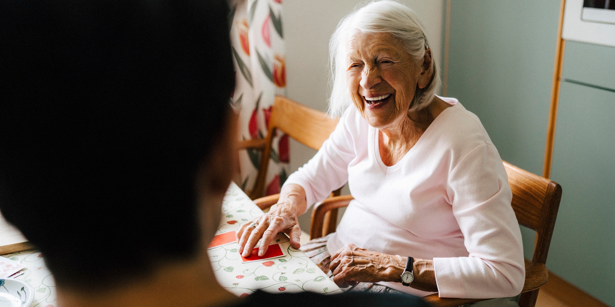 Äldre kvinna sitter ned vid bord och ler mot en ung hemtjänstanställd som står med ryggen mot kameran.