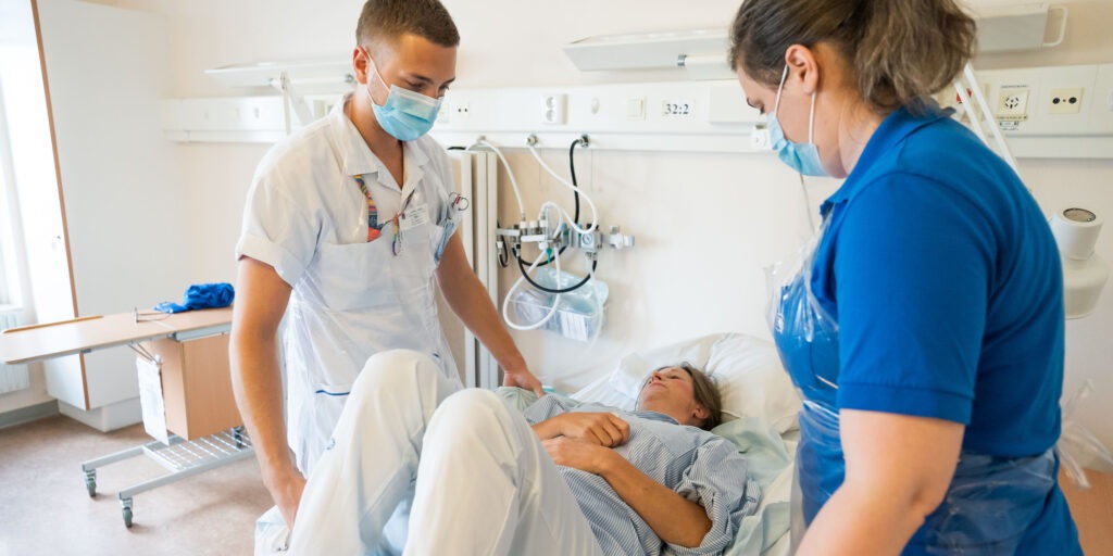Vårdpersonal hjälper patient i säng, tema säkra personförflyttningar.