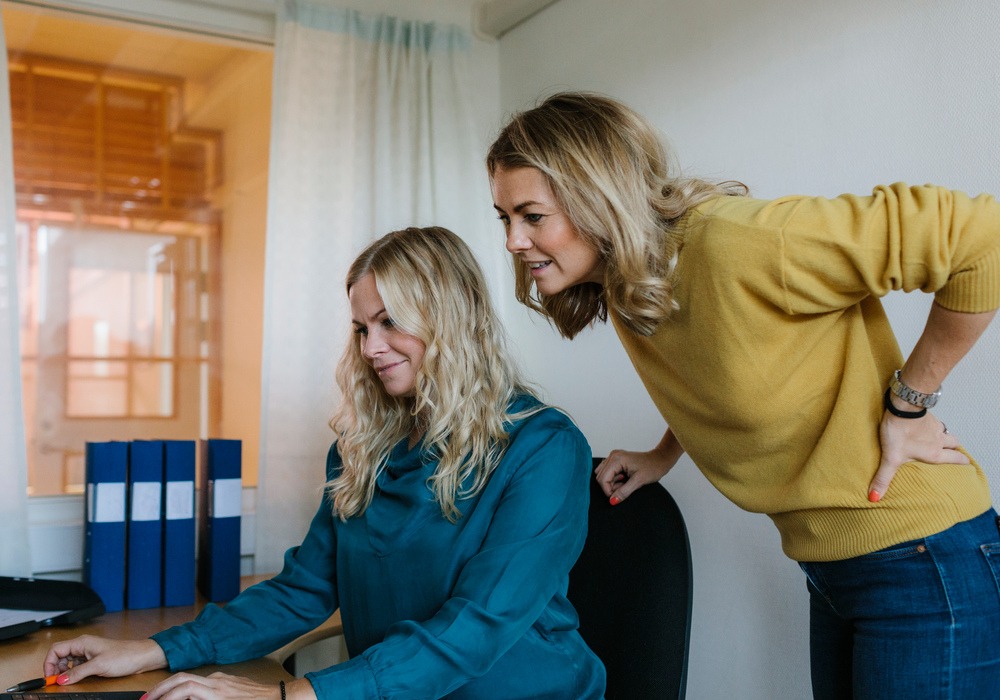 Två kvinnor vid ett skrivbord i kontorsrum - en sitter, en står.