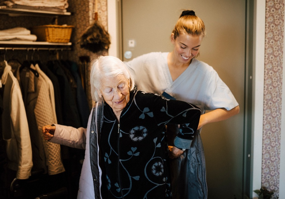 En yngre kvinna i hemtjänstkläder hjälper en äldre kvinna på med kappan. De står i en hall.