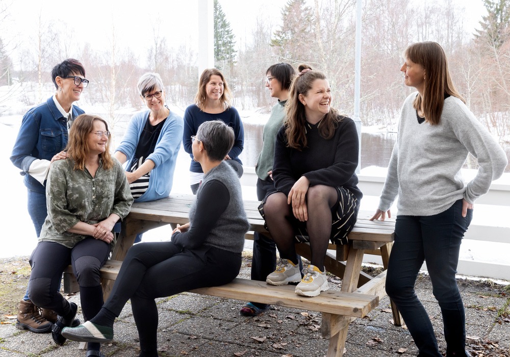 en grupp kvinnor står och sitter runt ett utomhusbord i trä. De tittar på varandra