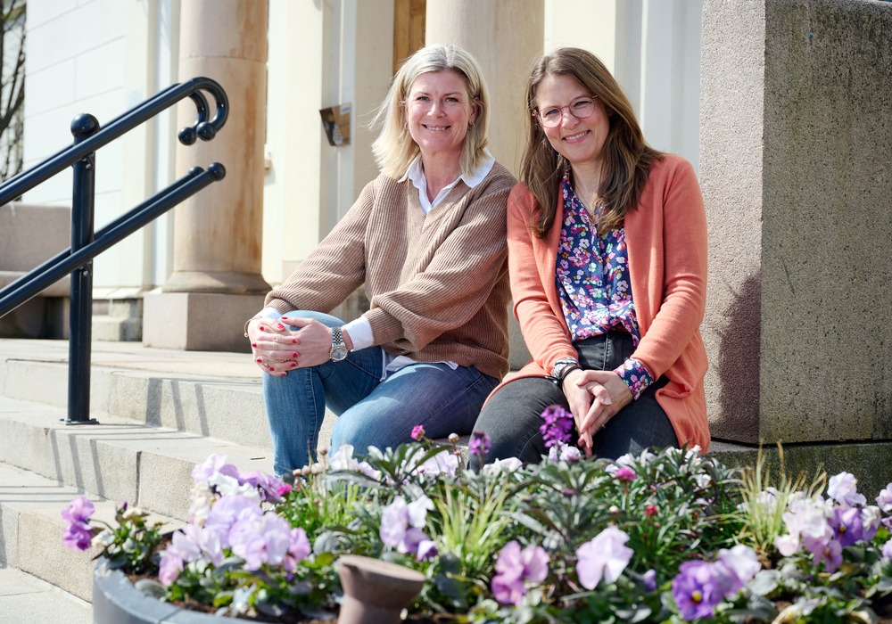 Anna Yström och Petra Hultqvist sitter på en trappa utanför stadshuset, med blommor i förgrunden