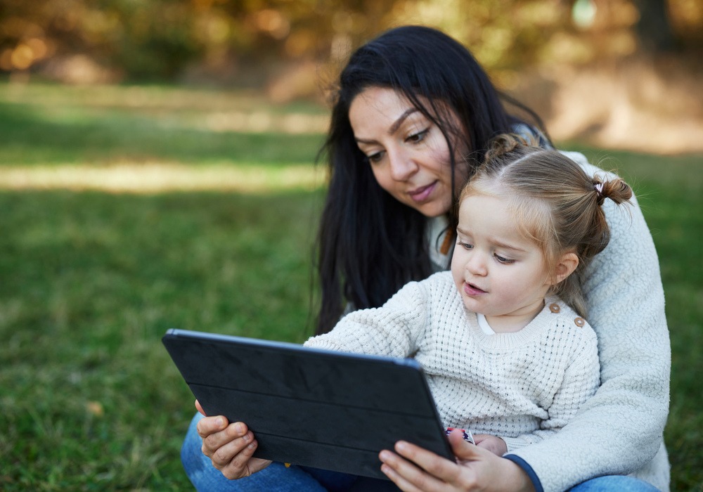 Mamma och barn sitter i park och ser på en Ipad tillsammans