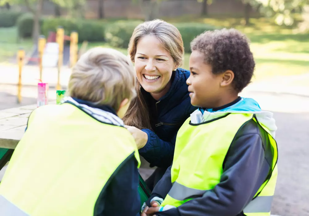Närbild å leende förskollärare med två förskolebarn i gula västar. Utomhus i förskolemiljö.