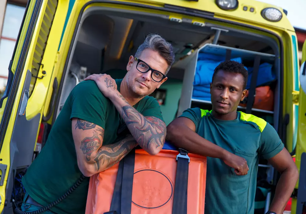 Två män från räddningstjänsten utanför en ambulans. Tittar in i kameran.
