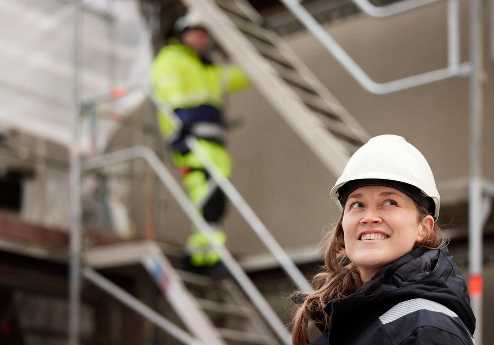 Porträtt på byggarbetare med hjälm, i bakgrunden byggarbetare går nerför trappa, tema arbetsskada.