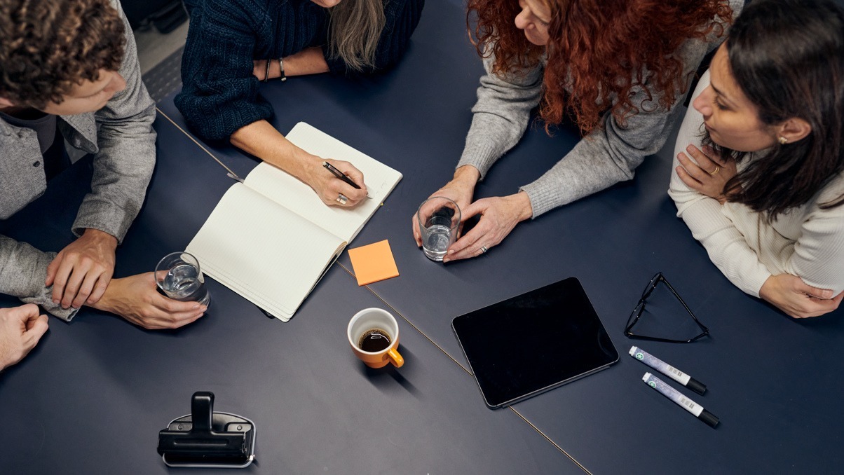 Bild ovanifrån av fyra personer som lutar sig över ett bord med skrivblock, pennor och post-it lappar på.