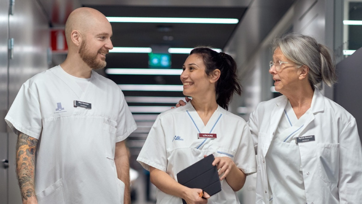 Tre leende kollegor som arbetar i sjukvården samtalar i en sjukhuskorridor.