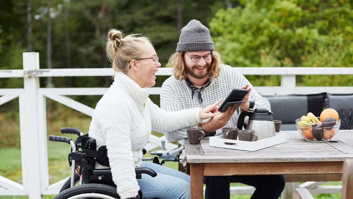 En man som jobbar som personlig assistent tittar på en läsplatta tillsammans med en ung kvinna i rullstol.