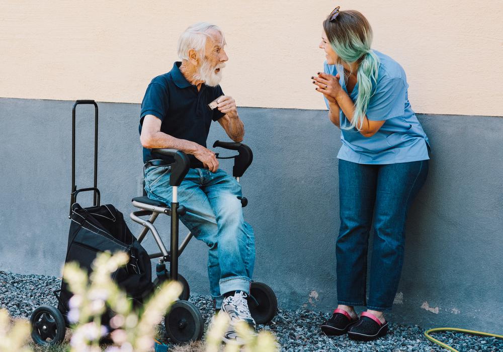 äldre man sitter på sin rullator i somrig utomhusmiljö, och pratar med en leende undersköterska som står lutad mot en vägg.