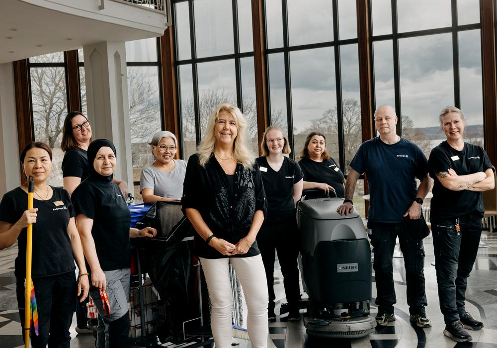 en stolt grupp städare står med maskiner och moppar  i ett stort rum med höga fönster