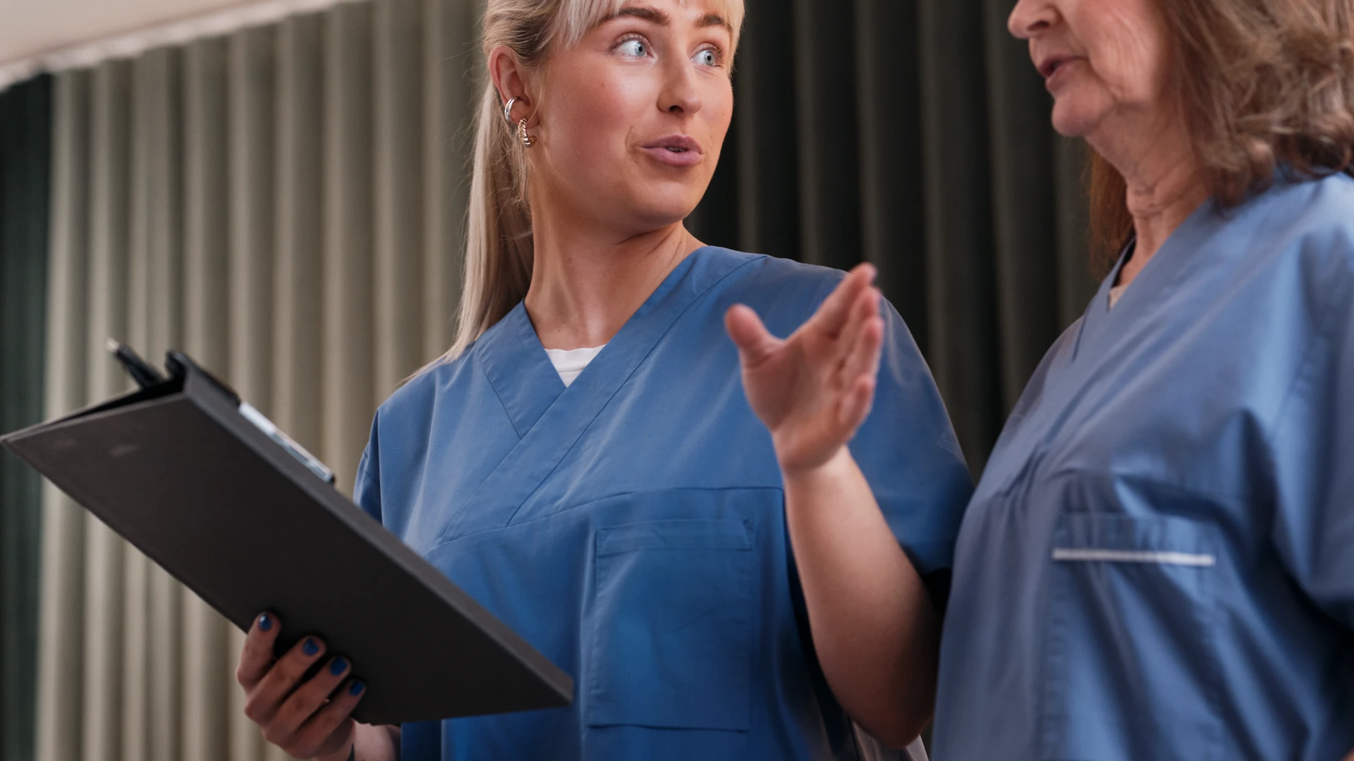 Två kvinnor i blå sjukhusskjortor samtalar i en korridor.