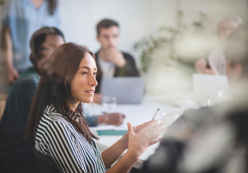 Grupp runt konferensbord, kvinna i fokus som talar.