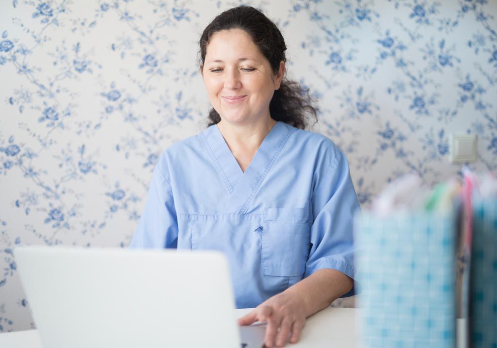 Sjuksköterskeklädd kvinna ler och sitter vid en dator i ett rum med ljusblå tapeter.