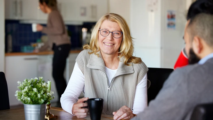En kvinna ser glad ut. Hon sitter med en kaffekopp i ett personalrum med kollegor.