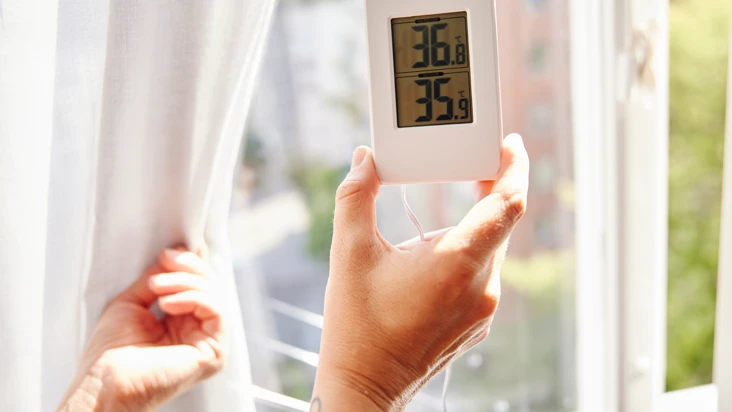 En hand håller upp en termometer mot ett somrigt fönster.